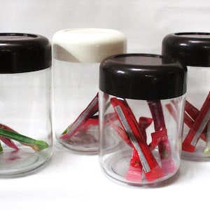 May include: Four clear glass jars with dark brown lids, each containing colorful items. One jar has a white lid. The jars hold red, green, and yellow packets. The jars are on a white surface.