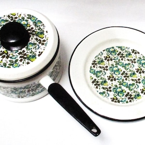 Puede incluir: Una cacerola de esmalte blanco con asa y tapa negras, y un plato a juego. Ambos presentan un estampado floral con hojas verdes, azules y amarillas. El juego está diseñado para cocinar y servir.