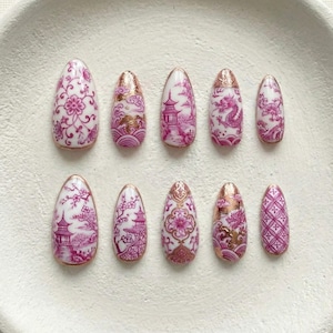 Könnte beinhalten: Ein Set aus zehn ovalen Press-on-Nägeln. Jeder Nagel hat eine weiße Basis mit rosa und goldfarbenen asiatisch inspirierten Designs, darunter Blumenmuster, Pagoden und Drachen. Die Nägel sind auf einer weißen, kreisförmigen Oberfläche angeordnet.