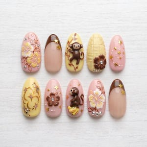 Op de afbeelding: Een set van tien kunstnagels met diverse ontwerpen, waaronder roze en gele hibiscusbloemen, bruine aapjes, gele bananen, geweven texturen en gouden steraccenten. De nagels zijn ovaal van vorm met glanzende afwerkingen en kleine steentjes.
