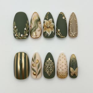 May include: A set of ten artificial nails in various shapes and designs. The nails feature olive green, gold, and beige colors with floral, striped, and abstract patterns. Some nails have 3D embellishments.