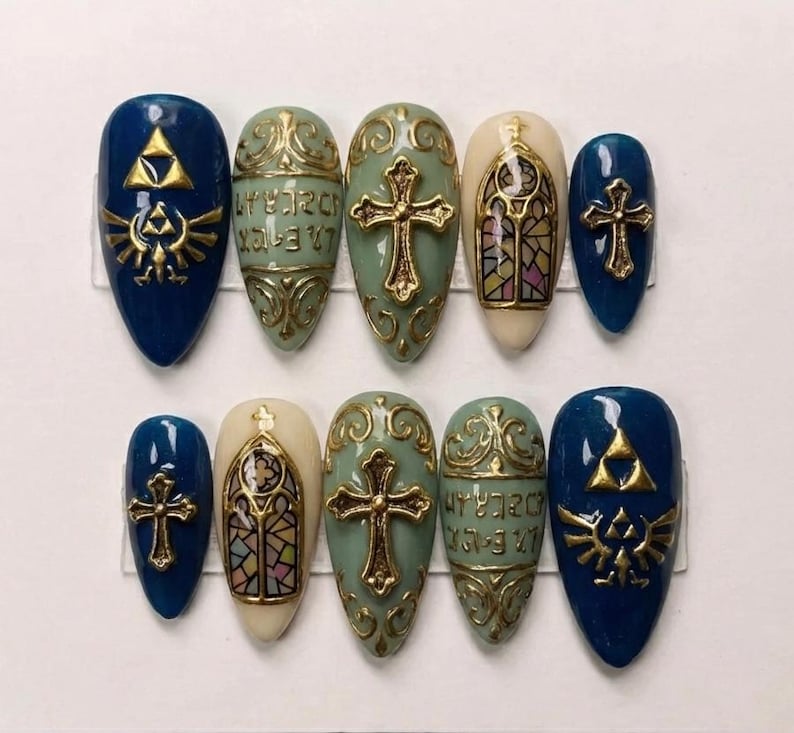 May include: A set of ten artificial nails in various designs. The nails feature a mix of colors, including blue, cream, and green, with gold accents. Designs include crosses, stained glass, and a gold emblem. The nails are oval-shaped.