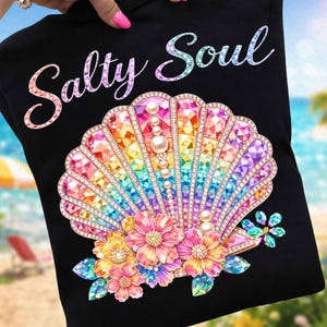 Puede incluir: Camiseta negra con la frase "Salty Soul". La camiseta presenta un diseño de concha marina colorida con gemas de arcoíris, perlas y detalles florales. El diseño es vibrante y llamativo, ideal para un look playero.