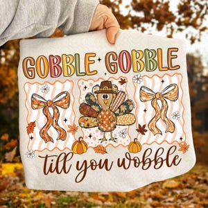 Puede incluir: Un cojín decorativo de color crema con temática de Acción de Gracias con las palabras "GOBBLE GOBBLE" en letras coloridas. El cojín presenta un pavo de dibujos animados con un sombrero de peregrino, lazos naranjas, calabazas y hojas de otoño. El texto "till you wobble" está escrito en cursiva.