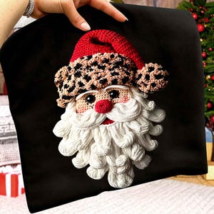 Peut inclure: Une housse de coussin noire avec un visage de Père Noël crocheté. Le Père Noël a un chapeau rouge avec une bande à imprimé léopard, une barbe blanche et un nez rouge. La housse est probablement pour la décoration de Noël.
