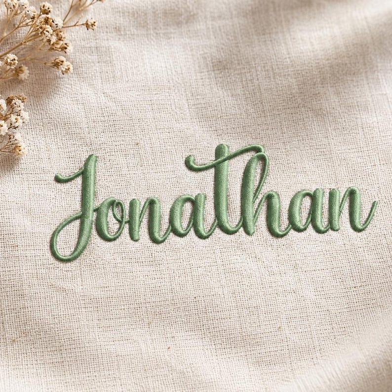 Op de afbeelding: De naam Jonathan is geborduurd in een saliegroen cursief lettertype op een lichtbeige, gestructureerde stoffen achtergrond. Een klein bosje gedroogde witte bloemen is zichtbaar in de linkerbovenhoek van het beeld voor een natuurlijk accent.