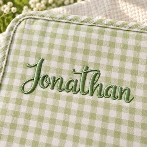 Op de afbeelding: Een lichtgroen en wit gingham stoffen item met de naam Jonathan geborduurd in een donkergroen cursief lettertype. De stof heeft een biesrand en een geweven textuur, met een klassiek ruitpatroon over het gehele oppervlak.