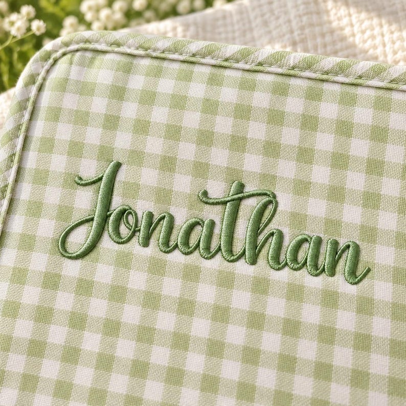 Op de afbeelding: Een lichtgroen en wit gingham stoffen item met de naam Jonathan geborduurd in een donkergroen cursief lettertype. De stof heeft een biesrand en een geweven textuur, met een klassiek ruitpatroon over het gehele oppervlak.