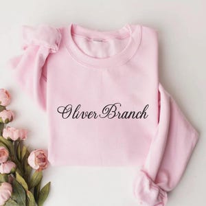 May include: A light pink long-sleeve sweatshirt featuring the text Oliver Branch printed in a black cursive script across the center of the chest. The garment has a crew neckline and ribbed cuffs on the sleeves, presented with pink flowers.