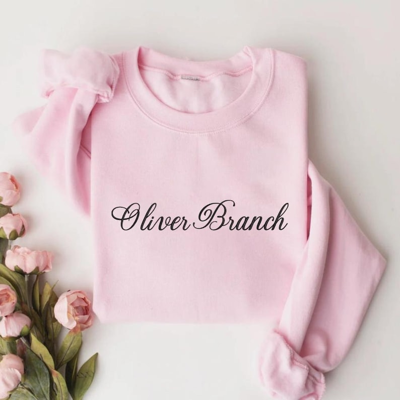 May include: A light pink long-sleeve sweatshirt featuring the text Oliver Branch printed in a black cursive script across the center of the chest. The garment has a crew neckline and ribbed cuffs on the sleeves, presented with pink flowers.