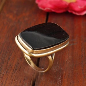 May include: A gold-toned ring with a large, square, black stone. The ring is set on a wooden surface with a pink flower in the background. The ring's design features a double band around the stone.