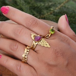 Peut inclure: Une main portant trois bagues en or. L'une d'elles est ornée d'une pierre précieuse rose foncé en forme de poire. Une autre bague présente une petite pierre verte ronde. La troisième bague est une alliance à motifs. Le fond est vert flou.