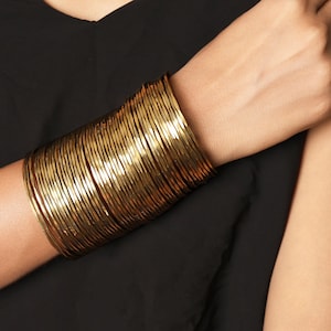 Puede incluir: Una pila de pulseras doradas en la muñeca. Las pulseras son finas y numerosas, creando una banda ancha y texturizada. La persona lleva una camiseta negra y su otro brazo es visible.