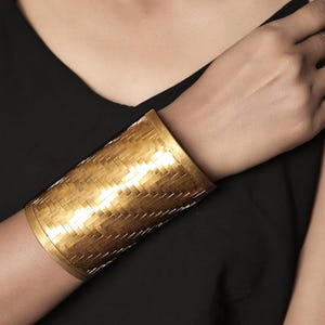 Puede incluir: Un brazalete ancho de color dorado con una textura tejida. El brazalete se lleva en la muñeca y el brazo de una persona que lleva una parte superior negra. El brazalete mide aproximadamente 7.6 cm de ancho.