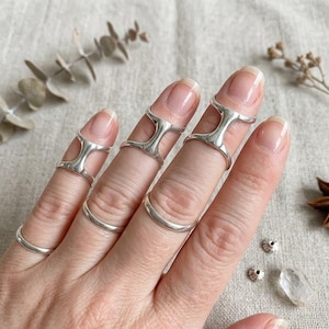 May include: A hand wearing several silver-colored finger rings. The rings have a unique design, with some covering the entire finger and others at the base. The rings are displayed on a neutral background.