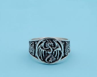 Può includere: Anello color argento con un elaborato disegno di drago al centro, racchiuso in un cerchio. Motivi a nodo celtico adornano i lati. L'anello è posto su uno sfondo azzurro chiaro. Il design è dettagliato.