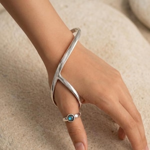 May include: A silver hand bracelet featuring a smooth, curved frame that wraps around the wrist and extends to a ring component. The ring is set with a small, round, iridescent blue gemstone that displays a subtle shimmer on the surface.