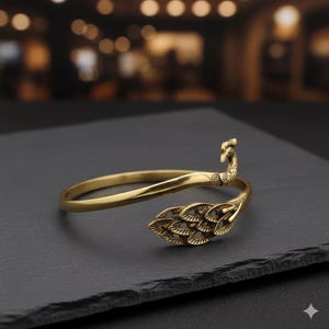 Może przedstawiać: Złota bransoletka na ramię z metalu. Bransoletka ma wzór pawia, z głową i piórami ogona jako elementami dekoracyjnymi. Bransoletka jest umieszczona na ciemnym tle.