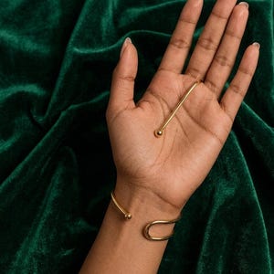 May include: A gold-toned metal cuff bracelet featuring a smooth, open-ended design with a rounded ball at each terminal end. The bracelet has a curved, U-shaped structure that wraps around the wrist, showcasing a polished, reflective surface finish.