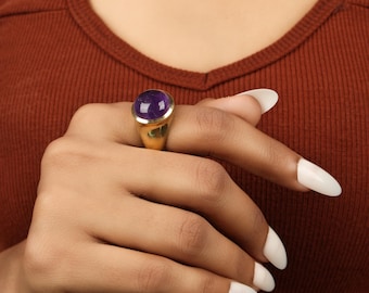 May include: A gold ring featuring a large, oval-shaped, deep purple gemstone. The ring has a thick band and a simple, elegant design. The gemstone is set in a bezel setting, which enhances its color and brilliance. The ring is worn on a finger.
