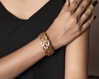 Puede incluir: Una pulsera dorada tejida. La pulsera está hecha de hebras de metal entrelazadas, creando un diseño texturizado y abierto. La joya se lleva en la muñeca de una persona, contra una parte superior negra.