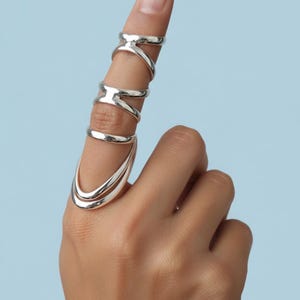 May include: A hand wearing multiple silver rings. The rings have unique designs, including an X-shape, a band, and a double-banded design. The rings are on the index finger, with a light blue background.