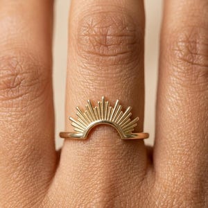 Puede incluir: Un anillo dorado con un diseño de rayos de sol. El anillo tiene una banda delgada, y el sol es el punto focal, con rayos que se extienden hacia arriba. El anillo se lleva en un dedo, mostrando sus delicados detalles y cálido tono metálico.