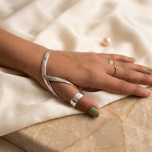 Könnte beinhalten: Ein poliertes Handarmband aus Silber mit V-förmigem Design, das ein Armband mit einem Daumenring verbindet. Das glatte, metallische Stück umschließt den Handrücken und bildet eine durchgehende Struktur, die um den Daumen herum befestigt ist.
