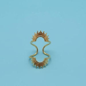 May include: A gold-colored ring with a sunburst design. The ring features two sunburst motifs, one at the top and one at the bottom, connected by curved bands. The ring is set against a light blue background.