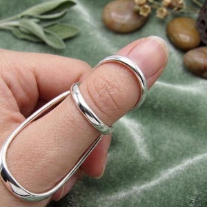 May include: A silver-colored metal finger splint featuring a smooth, polished finish. The device consists of two connected loops, with a smaller circular band encircling the base of the finger and a larger, elongated oval loop extending outward.