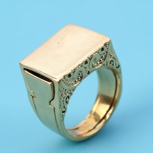 May include: A gold-colored signet ring with a rectangular top and ornate scrollwork on the sides. The ring has a hidden compartment and is set against a light blue background.