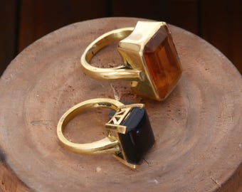 May include: Two gold-colored rings with large, rectangular gemstones. One ring features a dark amber-colored stone, while the other has a black stone. The rings are displayed on a wooden surface.