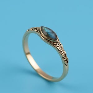 May include: A gold-colored ring with a marquise-cut, blue gemstone. The band features an ornate, filigree design. The ring is set against a solid blue background.