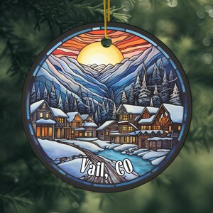May include: A round Christmas ornament with a stained glass design of a snowy mountain village at sunset. The ornament features a blue sky, white snow, and a yellow sun. The text "Vail, CO" is printed on the bottom of the ornament.