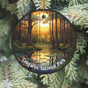 May include: A round, ceramic ornament featuring a stylized illustration of a forest scene with a river flowing through it. The ornament is decorated with a sunset and the text "Congaree National Park".