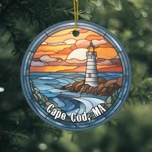 May include: Round glass ornament with a stained glass design of a lighthouse on a rocky shore at sunset. The text "Cape Cod, MA" is printed on the bottom of the ornament.