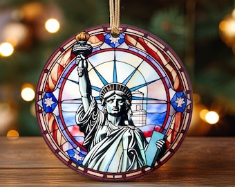 May include: A round Christmas ornament featuring a stained glass design of the Statue of Liberty. The ornament is decorated with red, white, and blue colors, reminiscent of the American flag.