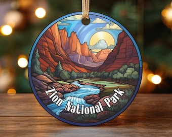 May include: A round, colorful Christmas ornament featuring a scenic view of Zion National Park. The ornament depicts a river flowing through a canyon with red rock cliffs and a bright yellow sun in the sky. The text "Zion National Park" is written in a curved arc at the bottom of the ornament.