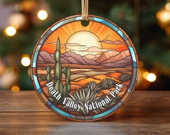 May include: A round, colorful Christmas ornament featuring a stained glass style depiction of a desert landscape with mountains, a setting sun, and cacti. The text "Death Valley National Park" is written in a blue arch at the bottom of the ornament.