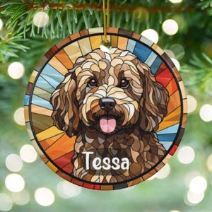 May include: A round ceramic ornament with a stained glass design featuring a brown and white dog with the name "Tessa" written below. The ornament has a colorful background with shades of blue, orange, yellow, and red.