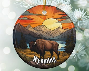 May include: Round ceramic ornament featuring a bison in a landscape with mountains, a lake, and a sunset. The word "Wyoming" is printed in white. The ornament has a gold string for hanging.