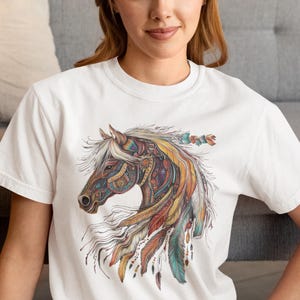 May include: White t-shirt with a colorful graphic of a horse's head with feathers and intricate designs.