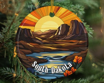 May include: A round ceramic ornament featuring a landscape design with a sunset, mountains, and a river. The ornament has the words "South Dakota" in white script. The design includes shades of orange, brown, and blue, with a gold hanging string.