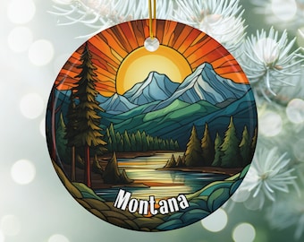 May include: Round ceramic ornament featuring a landscape scene with a sunset, mountains, and a lake. The ornament has a gold string for hanging and the word "Montana" is printed on the bottom.