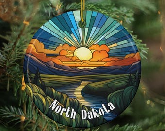 May include: A round Christmas ornament featuring a stained-glass style landscape of North Dakota. The design includes a sunset with orange and yellow hues, a river, and the text "North Dakota" in white.