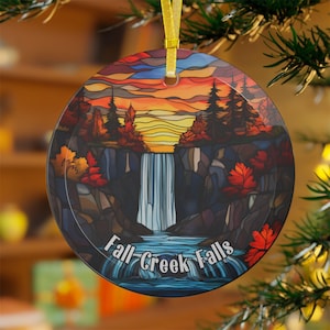 May include: Circular ceramic ornament with a stained-glass effect depicting a waterfall landscape. The design includes a sunset, trees, and the text "Fall Creek Falls". It is suspended by a gold ribbon.
