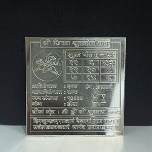 May include: A silver square plaque with engraved text and symbols. The plaque features a central grid, a star, and a circular image of a figure. The text is in a foreign script, with some English characters. The overall design suggests a religious or spiritual theme.