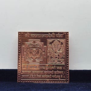 May include: A square copper-coloured metal plate featuring an engraved geometric yantra pattern on the left and a detailed depiction of a deity on the right. The top and bottom sections contain Sanskrit text inscriptions, all framed by a dotted border.