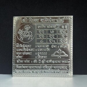 May include: A silver-colored, square religious plaque with intricate engravings. The plaque features detailed text and symbols, including a depiction of a figure riding an animal, and a grid of numbers. The text is in a foreign language.