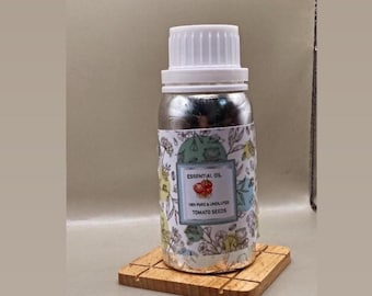 May include: A silver-colored bottle with a white screw-top cap, featuring a label with a floral pattern and text that reads ESSENTIAL OIL, 100% PURE & UNDILUTED, TOMATO SEEDS. The bottle is resting on a square, light brown wooden coaster.